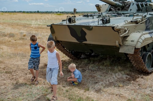 Preview: Three Children In Bright Clothes Are Sitting In Shade Under Modern Battle Tank. Children Play Until