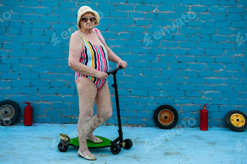 Preview: Elderly Woman In Striped Swimsuit, Sunglasses And Elegant Hat Riding Scooter Against Brick Wall