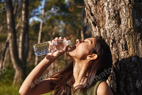Preview: Parched Young Lady Having A Drink Of Water While Siting On Natures Floor And Leaning Against A Tree