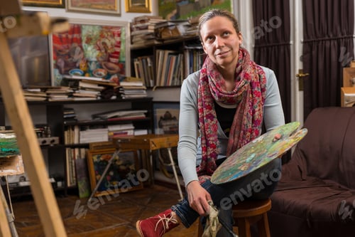 Preview: Thoughtful Female Artist Sitting In A Gallery Holding A Colorful Artists Palette And Paintbrush In