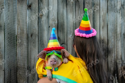 Preview: Eccentric Slim Woman With A Hat, Long Hair And Glasses Plays With A Funny Red Piglet In Hat For