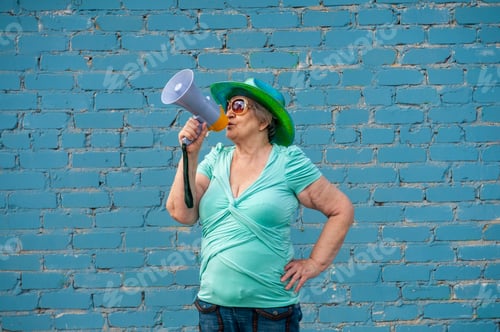 Preview: Elderly Grandmother In Turquoise Sweater And Cowboy Hat Stands With Megaphone Against Gray Brick