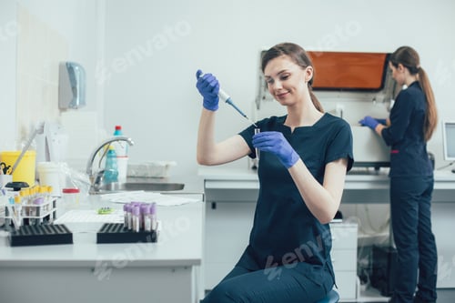 Preview: Cheerful Lab Assistant Sitting At The Table With A Test Tube In Her Hand And Smiling While Using A