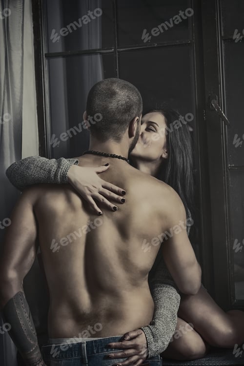 Preview: Enamored Brunette Woman In A Knitted Dress Sits On The Windowsill And Kissing Her Husband With Bare