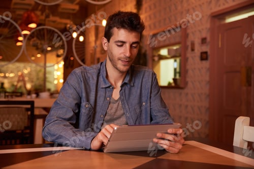 Preview: Handsome Young Man Wearing A Denim Shirt Is Sitting At A Table In A Restaurant With A Tablet That