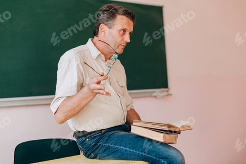 Preview: Middle Age Teacher Holding A Glasses And Sitting With Open Textbook.