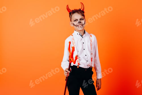 Preview: Devil Boy Standing Half-Turn Against An Orange Background In Masquerade Makeup. Halloween Concept