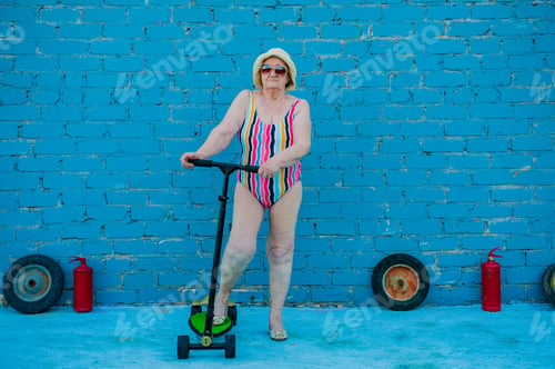 Preview: Elderly Woman In Striped Swimsuit, Sunglasses And Elegant Hat Riding Scooter Against Brick Wall