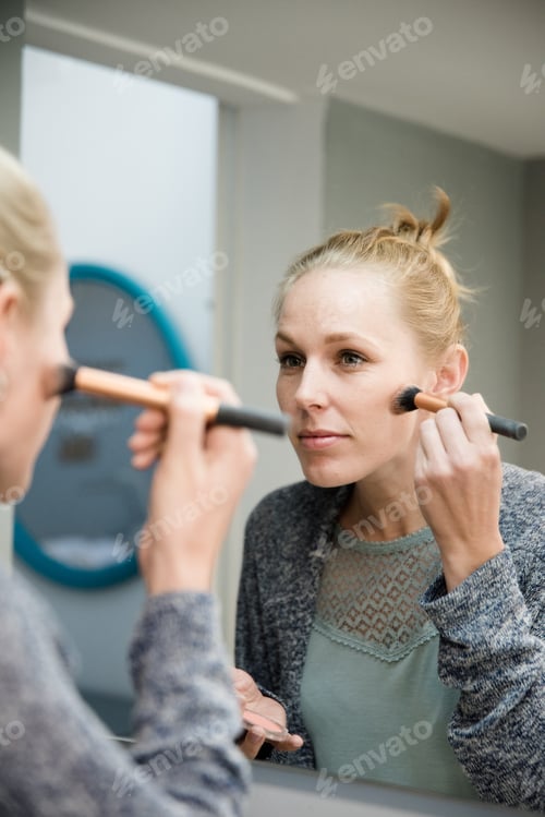 Preview: Woman Applying Makeup To Her Face As She Stands In Front Of A Mirror Wearing A Grey Jersey And Her