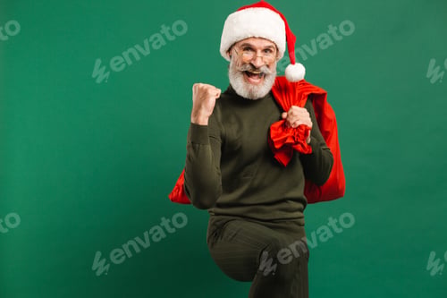 Preview: Happy Bearded Modern Santa Claus Holds A Red Bag And Shows A Fist Of Joy On A Green Background
