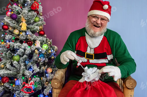 Preview: Elderly Man With Gray Beard And Glasses In Santa Claus Hat With Red Bag Filled With Presents Poses