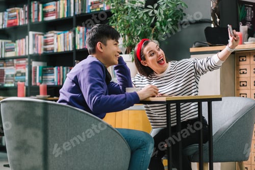 Preview: Beautiful Happy Couple Sitting In A Cafe In Comfortable Armchairs And Taking Selfies. Bookshelves