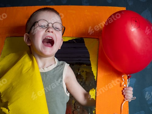 Preview: Funny Boy In Blue Clothes And Glasses Is Playing With Red Balloon Against Gray Wall Against