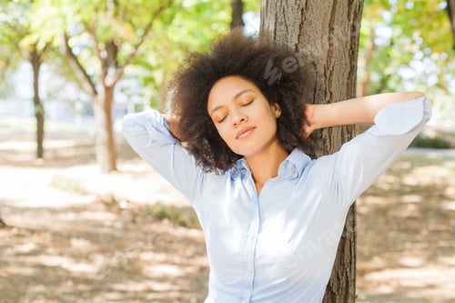Preview: Beautiful Smiling African Young Woman Dreaming With Closed Eyes In Nature, Leaning On Tree In Park