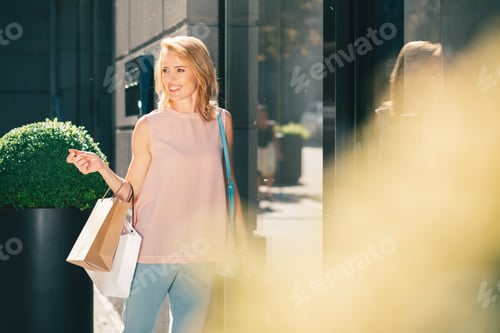Preview: Mirthful Young Woman Carrying Paper Bags And Smiling While Being Outdoors In Sunny Weather