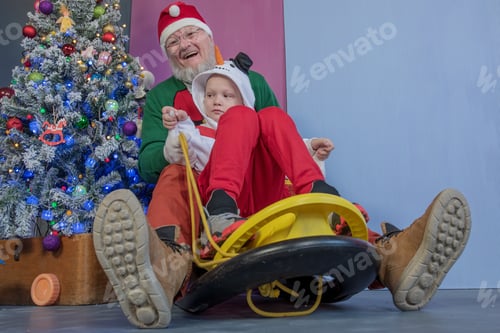 Preview: Elderly Man In Santa Claus Costume Is Rolling In Yellow Sled With Child Boy In Snowman Costume
