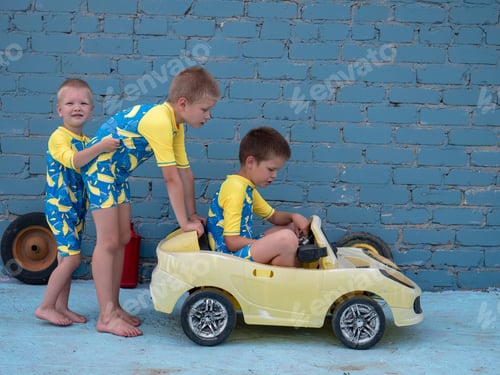Preview: Funny Children In Bathing Suits For Boys Roll Each Other In Yellow Car Against Background Of Brick