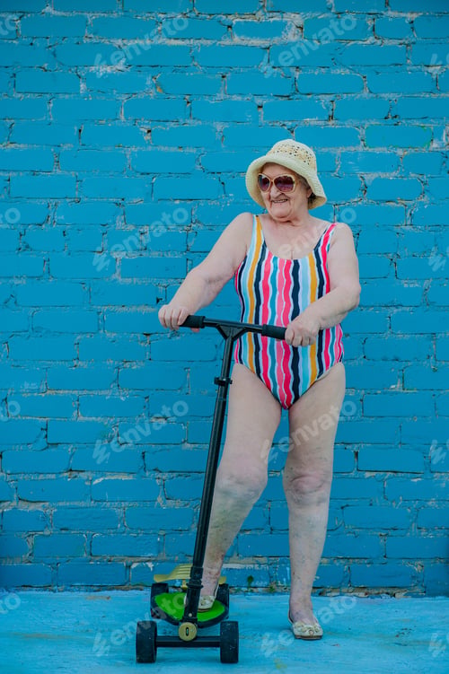 Preview: Elderly Woman In Striped Swimsuit, Sunglasses And Elegant Hat Riding Scooter Against Brick Wall
