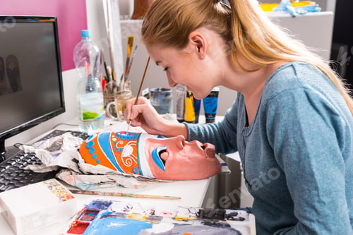 Preview: Close Up Of Young Artistic Woman Carefully Painting Colorful Details Onto Clay Mask At Computer Desk