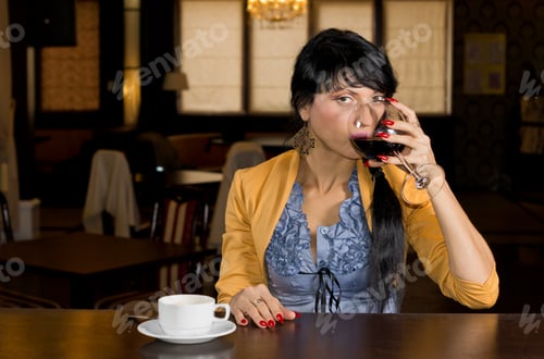 Preview: Attractive Young Woman Drinking Wine And Coffee At A Bar Counter Sipping From A Large Wineglass