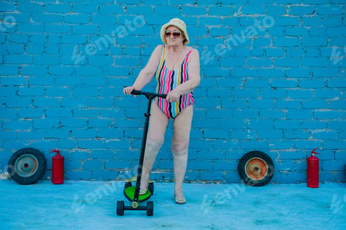 Preview: Elderly Woman In Striped Swimsuit, Sunglasses And Elegant Hat Riding Scooter Against Brick Wall