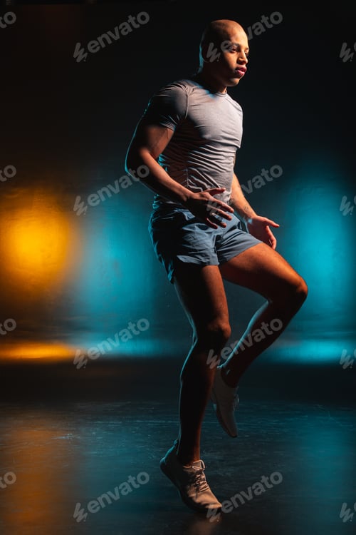 Preview: Bold Young Man In Shorts And A T-Shirt Looking Serious While Jogging. Orange Teal Light On The