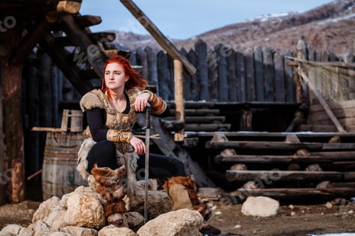 Preview: Red-Haired Woman Is A Viking. Against The Backdrop Of A Village Or An Ancient Settlement. In The