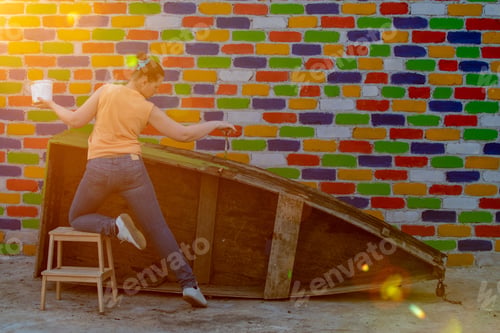 Preview: Young Slender Caucasian Woman Paints Wooden Boat Against Brick Wall Background. Brush, Container