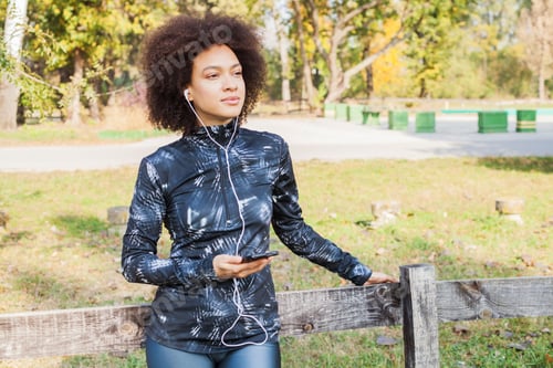 Preview: Sporty African American Woman Relax Listening Music After Training At Park, Leaning On Wooden Fence