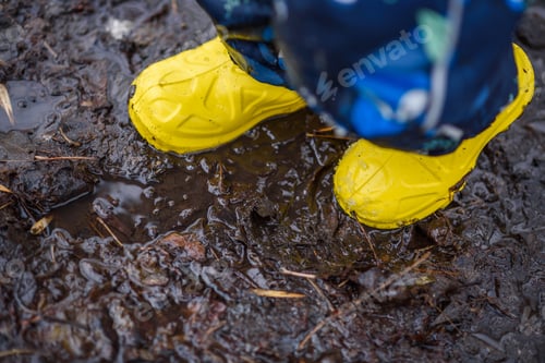 Preview: A Small Child In Yellow Rubber Boots Stands In The Mud