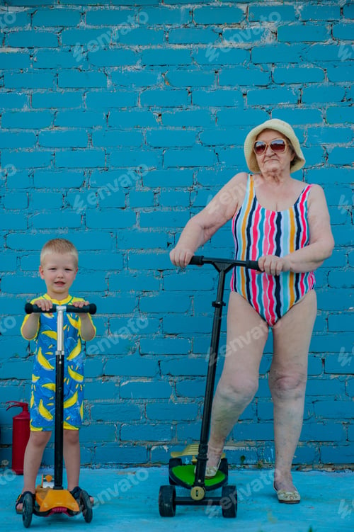 Preview: Elderly Woman In Striped Swimsuit, Sunglasses And Elegant Hat Riding Scooter Against Brick Wall
