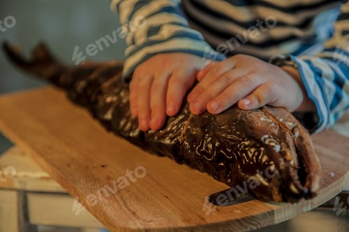 Preview: Little Blond Boy In Cook'S Hat Shows Big Smoked Fish. Child Tries Fish With Knife And Fork. Kid