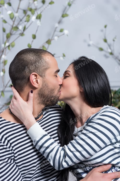 Preview: Charming International Couple In Striped Sweaters Kissing And Hugging While Sitting On The Bed In