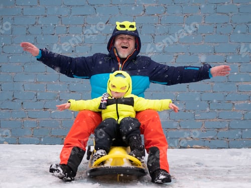 Preview: Dad And Child In Colorful Winter Clothes And Mask Riding Sleigh Along Gray Brick Wall In Snow. Man