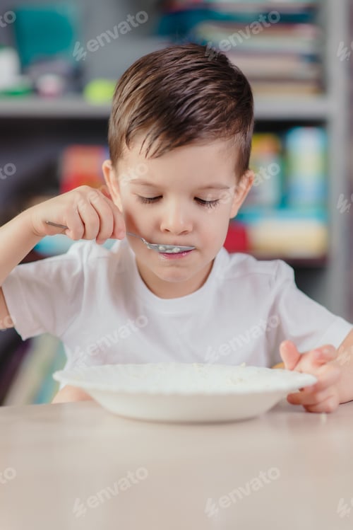 Visualização: Cute Baby Eats Milk Porridge. Beautiful Boy Has Breakfast Sitting At A Table In The Kitchen At Home.