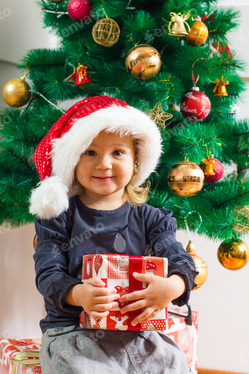 Preview: Happy Little Girl With Santa Hat Holding A Christmas Gift At Home.