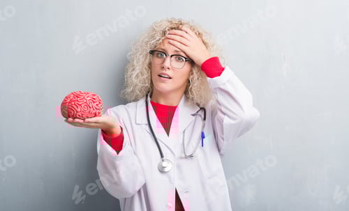 Preview: Young Blonde Doctor Woman Over Grunge Grey Wall Holding Brain Stressed With Hand On Head, Shocked