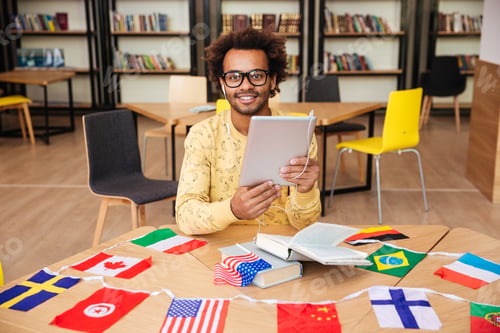 Preview: Happy African American Young Man In Glasses With Tablet And Flags Of Diffrent Countries Sitting In