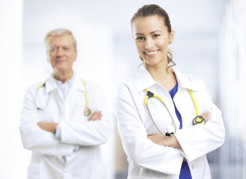 Preview: Smiling Young Female Doctor Standing With Arms Crossed In Hospital Ward. Her Colleague In The
