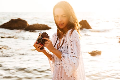 Preview: Pretty Brunette Woman In Light Summer Dress Posing Sideways On Beach With Retro Camera In Hands And