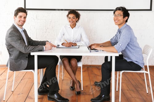 Preview: Group Of Smiling Young Multiethnic Businesspeople Sitting At The Desk And Looking At Camera