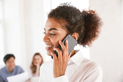 Preview: Close Up Of A Happy Young African Woman Talking On Mobile Phone With Talking Colleagues On A