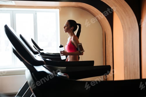 Preview: Profile Of Attractive Serious Young Sportswoman Walking On Treadmill And Drinking Water In Gym