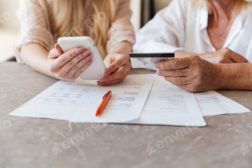 Preview: Cropped Photo Of Young Lady Sitting At Home With Her Grandmother Using Mobile Phone Holding Credit