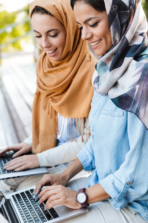 Preview: Photo Of Cheerful Islamic Girls Wearing Headscarfs Studying In Park And Using Silver Laptops