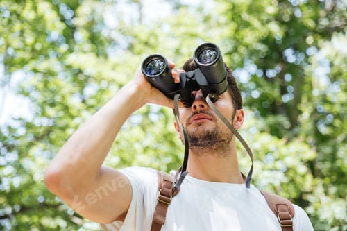 Preview: Concentrated Young Man With Backpack Looking Through Binoculars In Forest