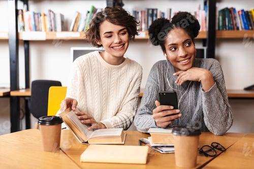 Preview: Two Cheerful Young Girls Students Studying At Library, Holding Mobile Phone,