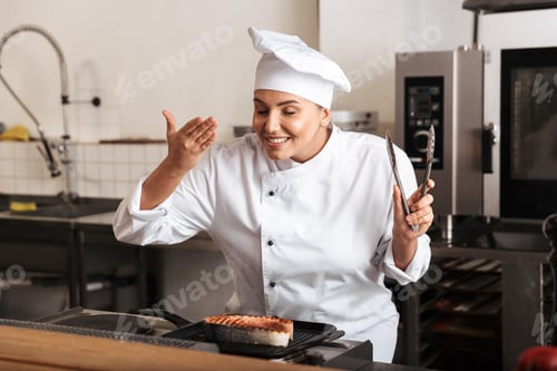 Preview: Smiling Woman Chef Cook Wearing Uniform Cooking Delicious Salmon Steak Standing At The Kitchen