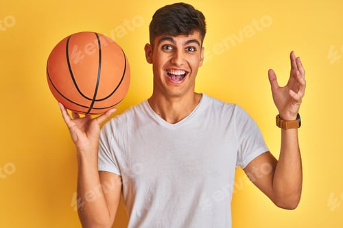 Preview: Young Indian Sportsman Holding Basketball Ball Standing Over Isolated Yellow Background Very Happy