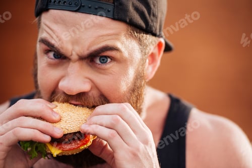Preview: Angry Irritated Young Man In Cap Eating Hamburger Outdoors